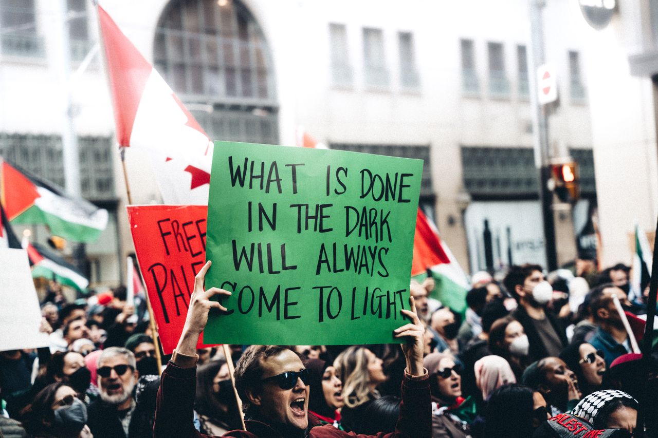A diverse crowd participates in a protest holding signs and flags, advocating for justice and visibility.
