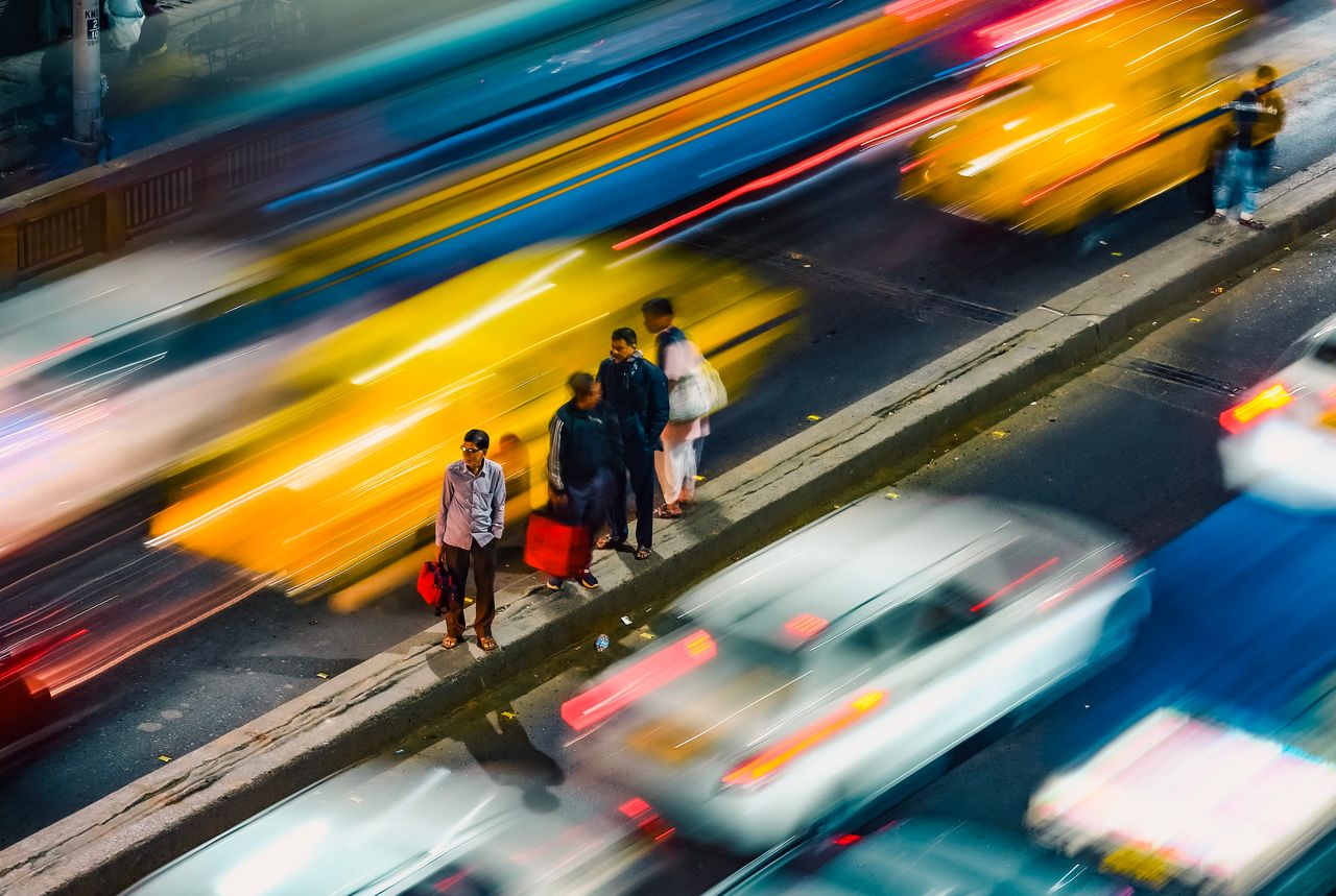 Blurred traffic rushes past pedestrians in Kolkata at night, capturing urban chaos and vibrant movement.