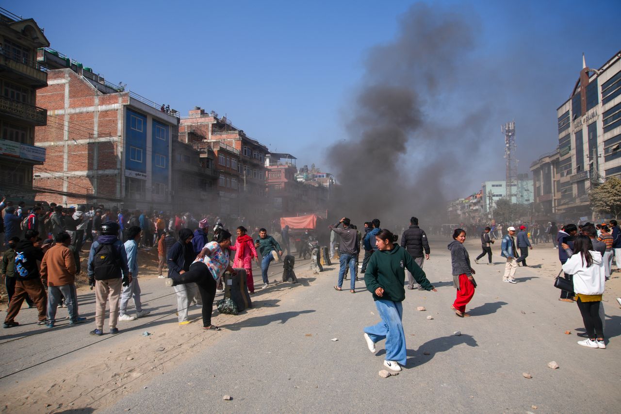 A large crowd gathers on a busy street as smoke rises into the sky. Urban setting, clear day.