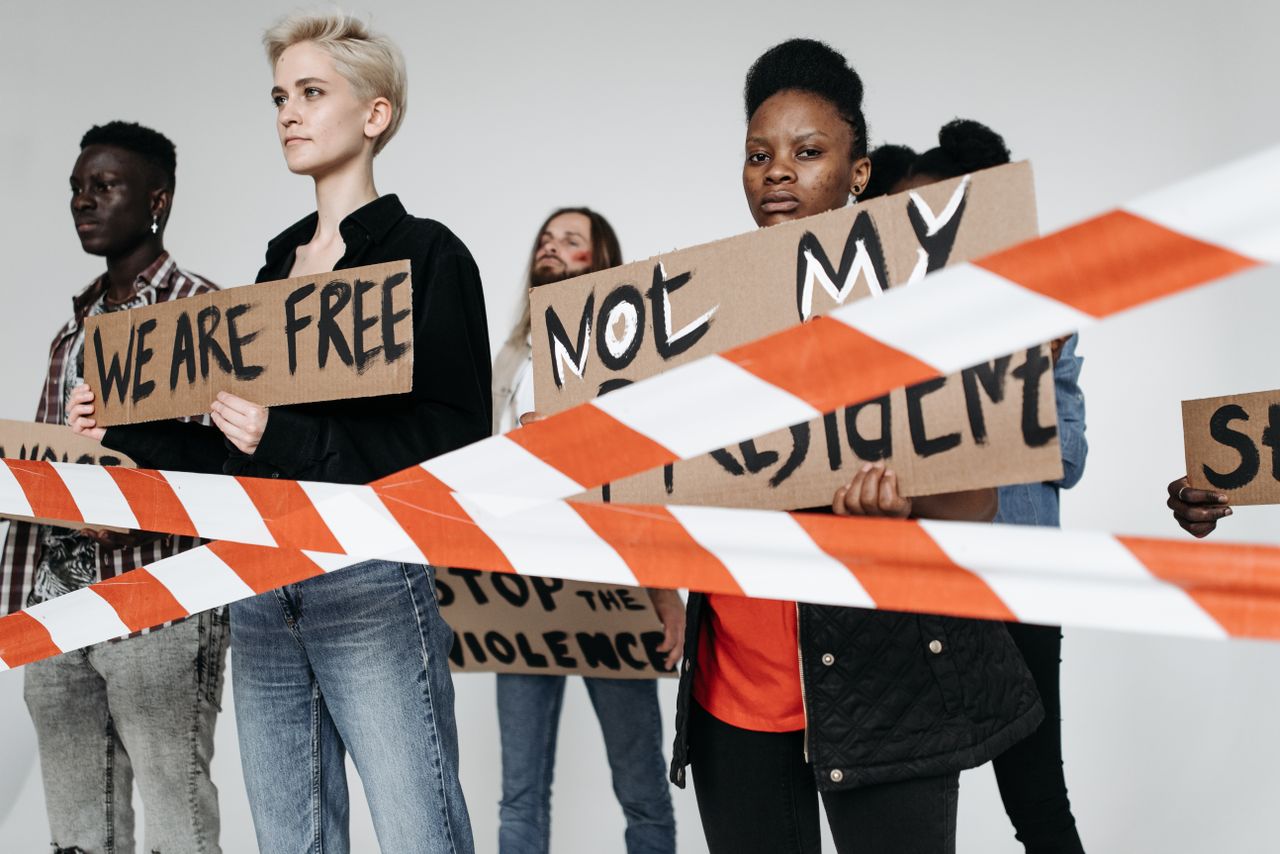 A diverse group of people holding protest signs indoors with police tape in the foreground, reflecting social activism.